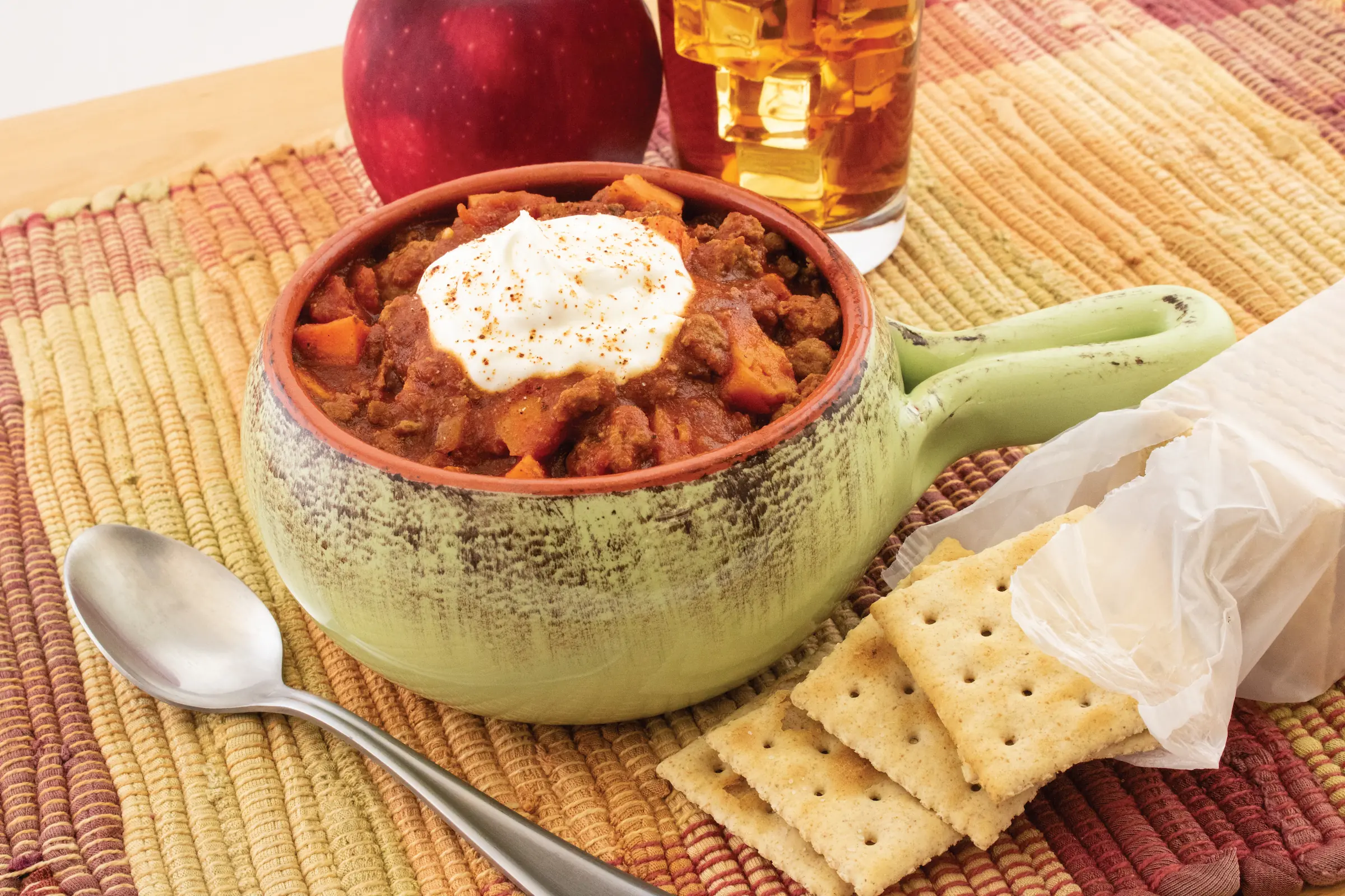 Chipotle turkey chili is prepared in a bowl with a handle, sitting atop a table with some saltine crackers on the side.